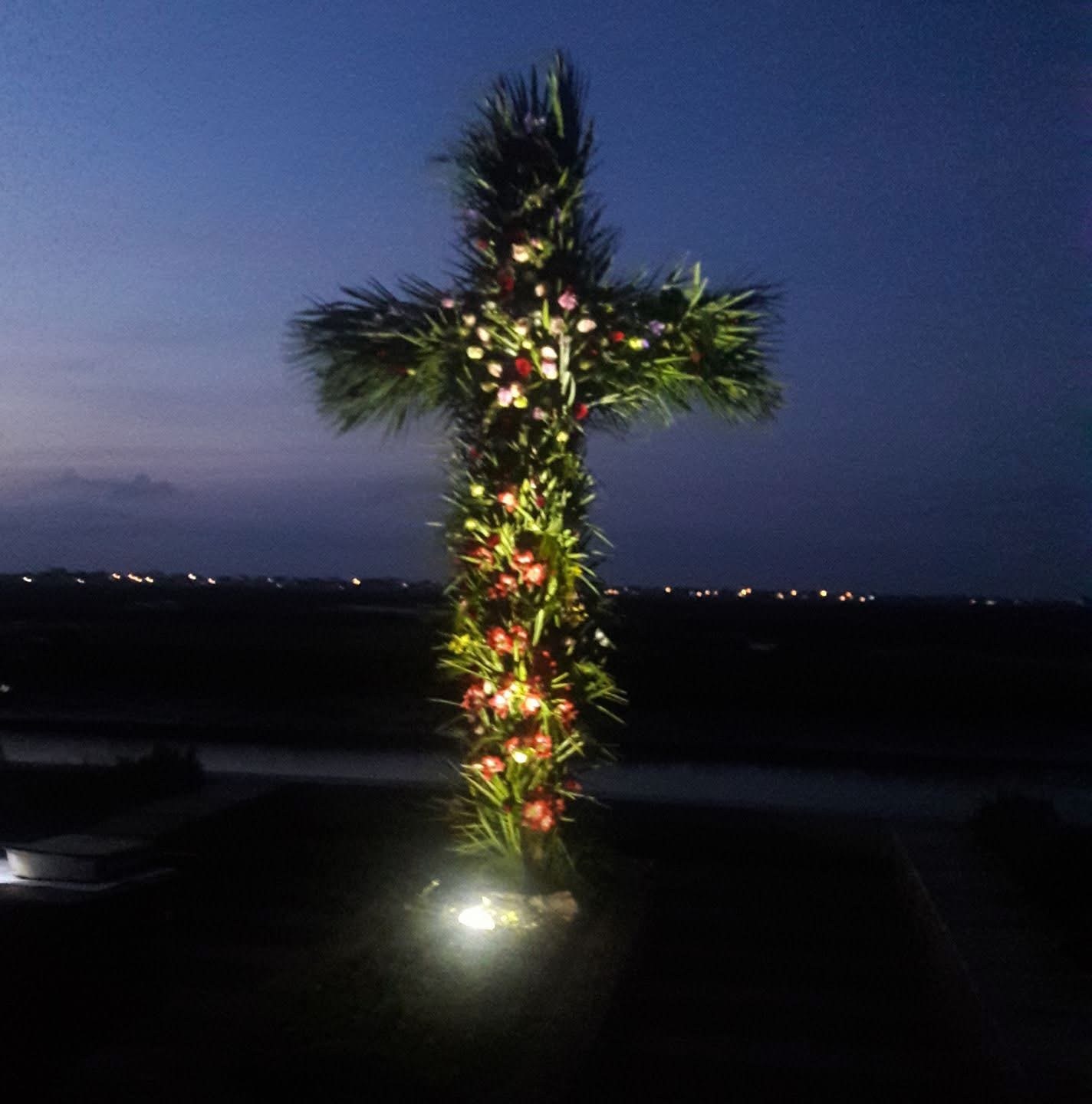 A cross of greenery and flowers, illuminated against the evening sky